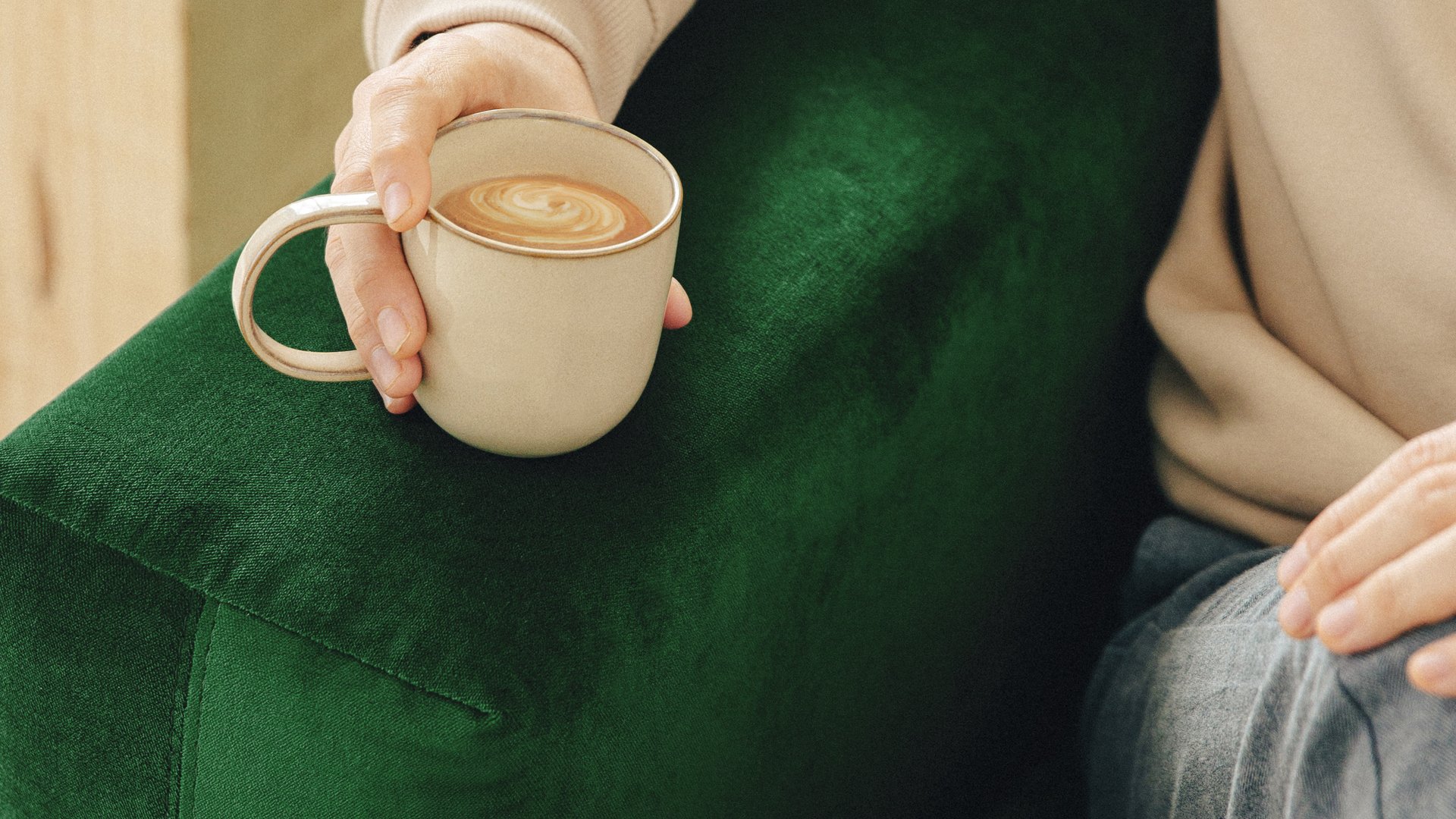 Lähikuva vihreästä samettikankaisesta sohvasta, keskellä kuvaa käsi joka pitelee kahvikuppia.