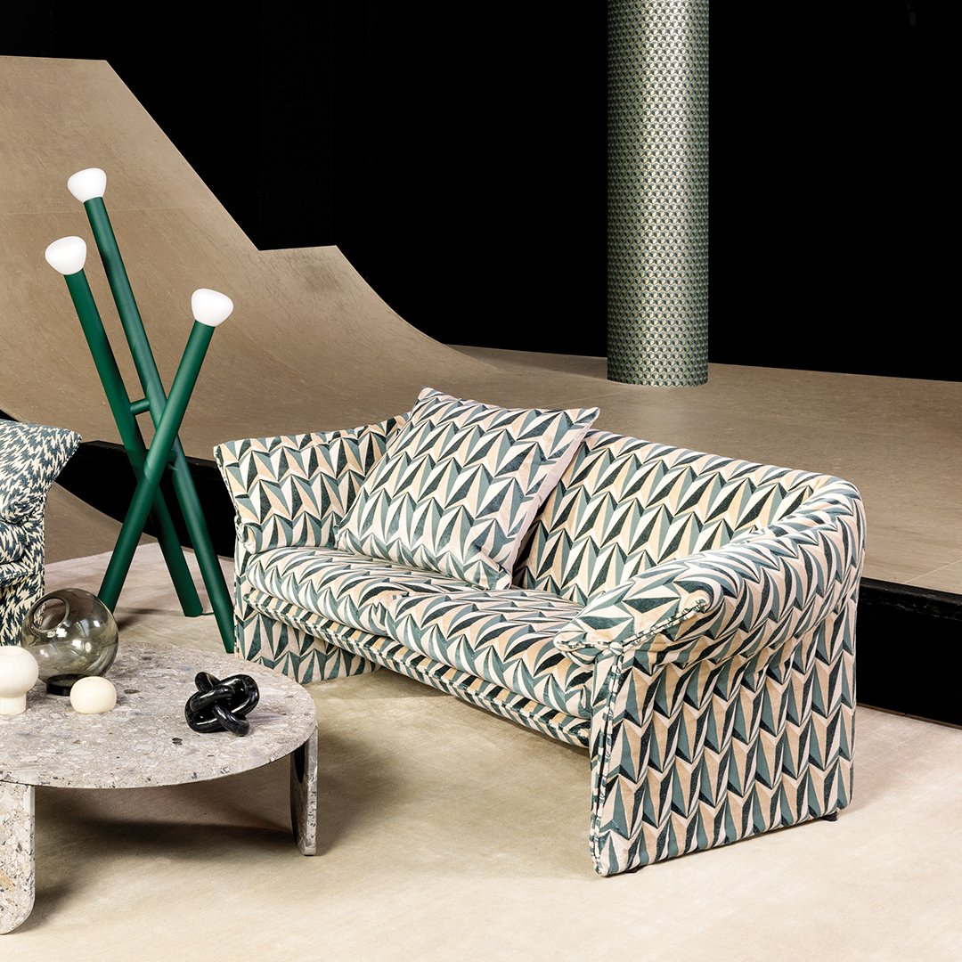 Keskellä graafinen kolmiokuvioitu sohva, jossa kiiltävä samettinen hohto. Marmorisella sohvapöydällä koriste-esineitä.