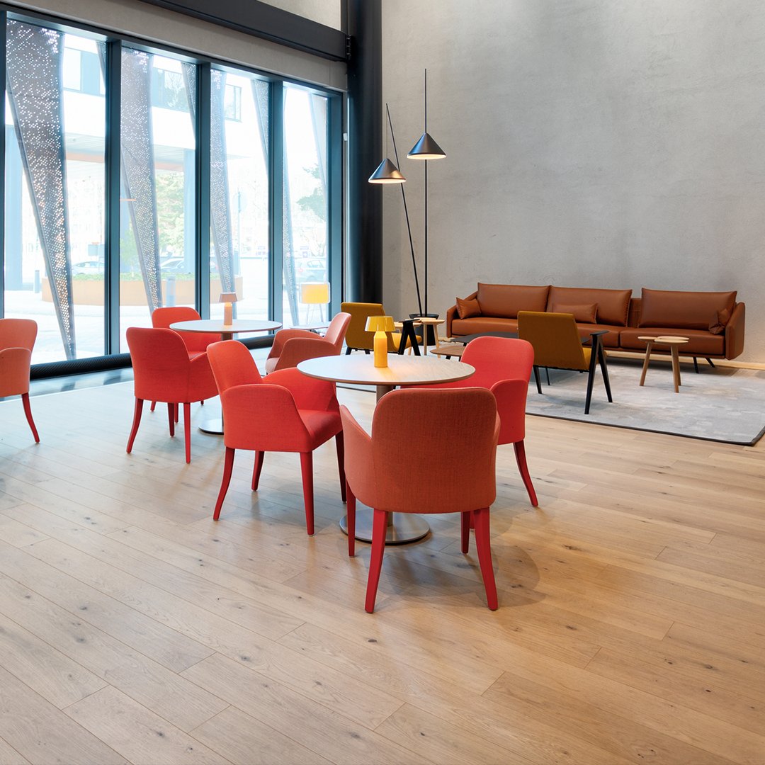Sisustuskuva Swing House-toimitalon aulatilasta, jossa leveälankkuinen Parky-puulattia, sekä punaisten tuolien pöytäryhmiä. Taustalla konjakinruskea nahkasohva.