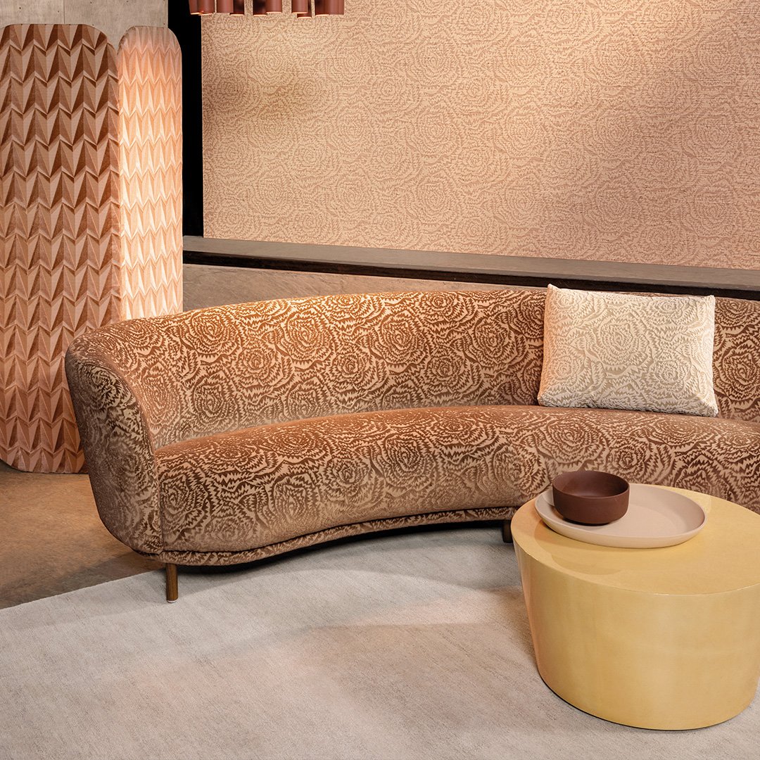 Kaarevanmuotoinen sohva, jossa persikanoranssi ruusukuvioinen verhoilu. Vieressä pyöreä keltainen sohvapöytä.